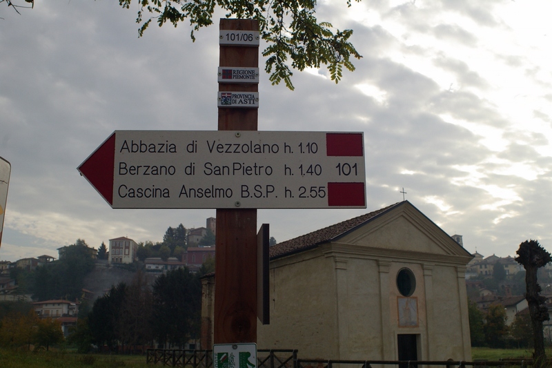 Cartello  indicatore del sentiero per la Canonica di Santa Maria di Vezzolano e sullo sfondo la Chiesa romanica di Sant Eusebio.